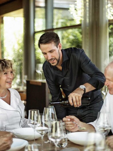 A group of four people is sitting at a table in a restaurant. A waiter is serving wine while the guests are engaging in pleasant conversation. | © SIGNUM BLANC communications