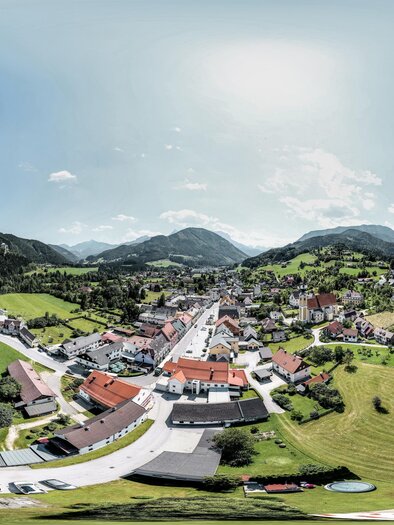 Eine malerische Ansicht eines kleinen Dorfes umgeben von Bergen und grünen Wiesen. Der Himmel ist teilweise bewölkt und der Ort strahlt eine ruhige Atmosphäre aus.