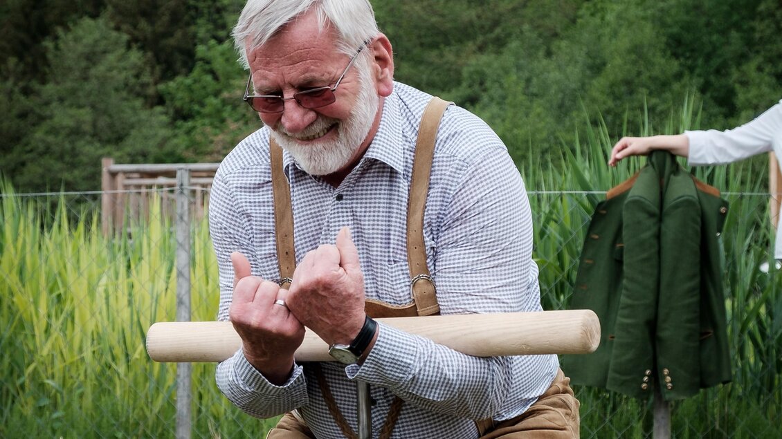 Ein älterer Mann in traditioneller Kleidung steht an einem Metallbehälter und benutzt eine Kurbel. Im Hintergrund sind grüne Wiesen und Bäume zu sehen. | © @Florian Jöbstl