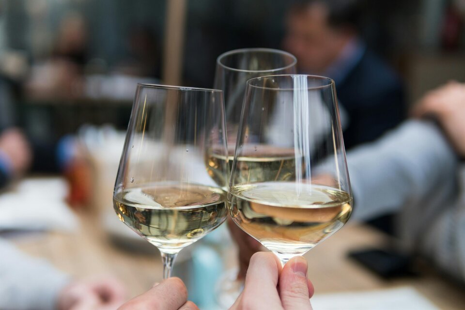 Zwei Gläser mit Weißwein stoßen an. Im Hintergrund sind verschwommene Menschen bei einem geselligen Treffen zu sehen. | © matthieu-joannon-unsplash