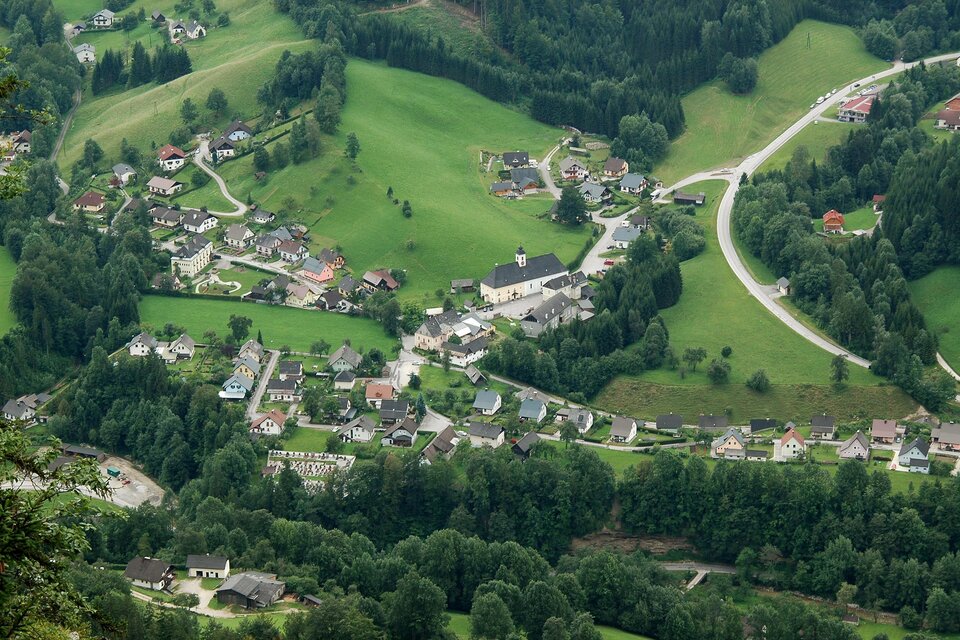 Eine malerische Landschaft mit einem kleinen Dorf, umgeben von grünen Wiesen und Wäldern. Die Straßen schlängeln sich durch die sanften Hügel.