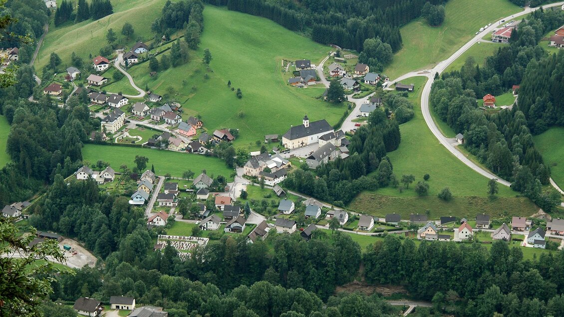 Eine malerische Landschaft mit einem kleinen Dorf, umgeben von grünen Wiesen und Wäldern. Die Straßen schlängeln sich durch die sanften Hügel.