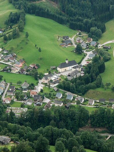 Eine malerische Landschaft mit einem kleinen Dorf, umgeben von grünen Wiesen und Wäldern. Die Straßen schlängeln sich durch die sanften Hügel.