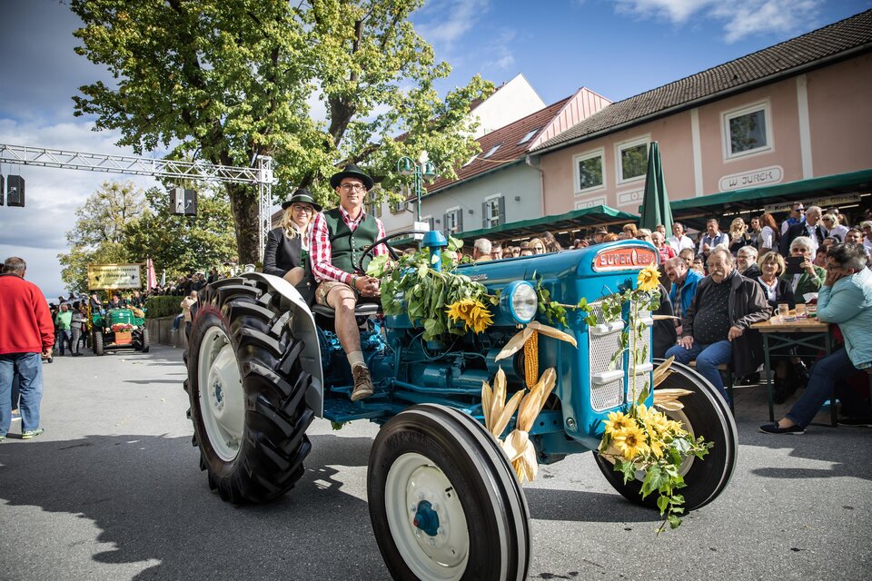 Ein festlich geschmückter Traktor fährt durch eine belebte Straße. Im Hintergrund stehen Zuschauer und feiern das Ereignis. | © Achromatic Photography