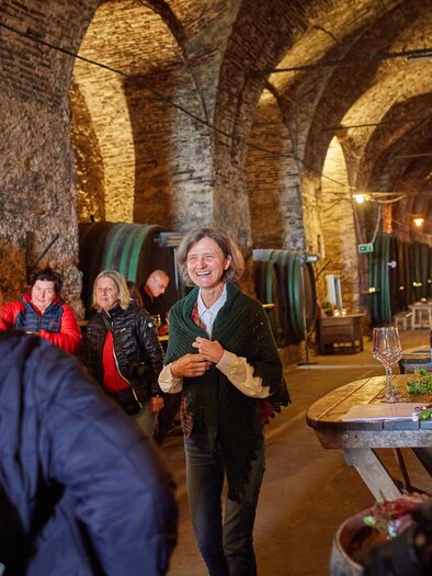 Eine Gruppe von Menschen in einem Weinkeller hört einer lächelnden Frau zu, die anscheinend führt. Auf einem Tisch sind Weingläser und Flaschen mit Wein zu sehen. | © Janez Kotar
