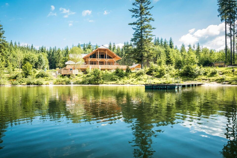 Am Ufer des ruhigen Ödensees steht die stilvolle Kohlröserlhütte umgeben von dichten Wäldern. Der Himmel ist klar und die Reflexion des Hauses im Wasser ist gut sichtbar. | © Die Abbilderei Dieter Sajovic