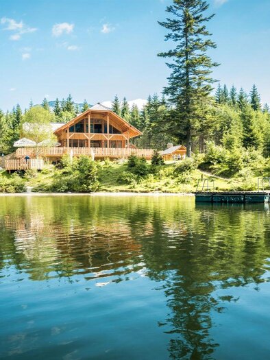 On the shore of the calm Ödensee lake stands the stylish Kohlröserlhütte hut, surrounded by dense forests. The sky is clear and the reflection of the house in the water is clearly visible. | © Die Abbilderei Dieter Sajovic