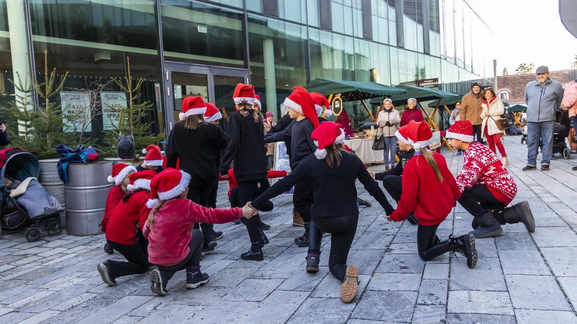 Eine Gruppe von Kindern in Weihnachtsmützen tanzt draußen vor einem modernen Gebäude. Im Hintergrund sind Zuschauer und Weihnachtsdekoration zu sehen.