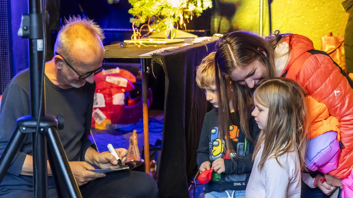 Ein Mann sitzt an einem Tisch und signiert etwas. Zwei Kinder und eine Frau schauen neugierig zu.