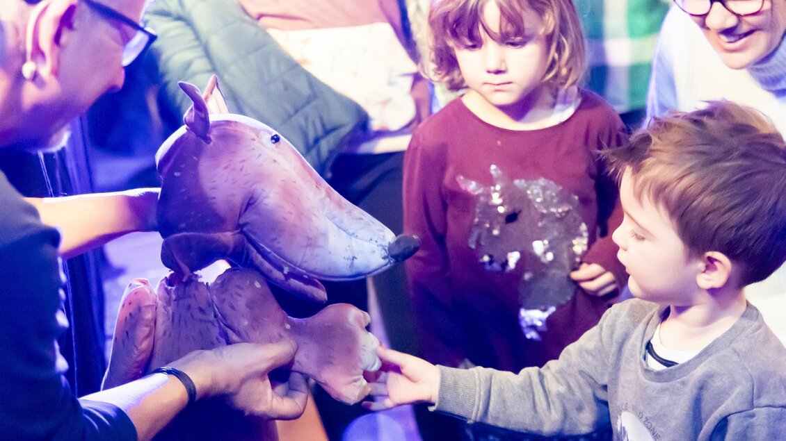 Ein Puppenspieler zeigt Kindern eine Figur eines Hundes. Die Kinder lächeln und scheinen begeistert zu sein.