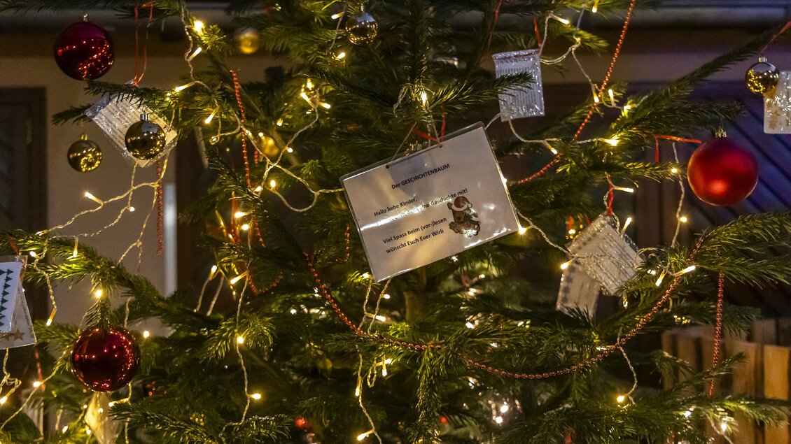Ein festlich geschmückter Weihnachtsbaum mit roten Kugeln und Lichtern. Veilchen und Geschenkkarten sind ebenfalls sichtbar.
