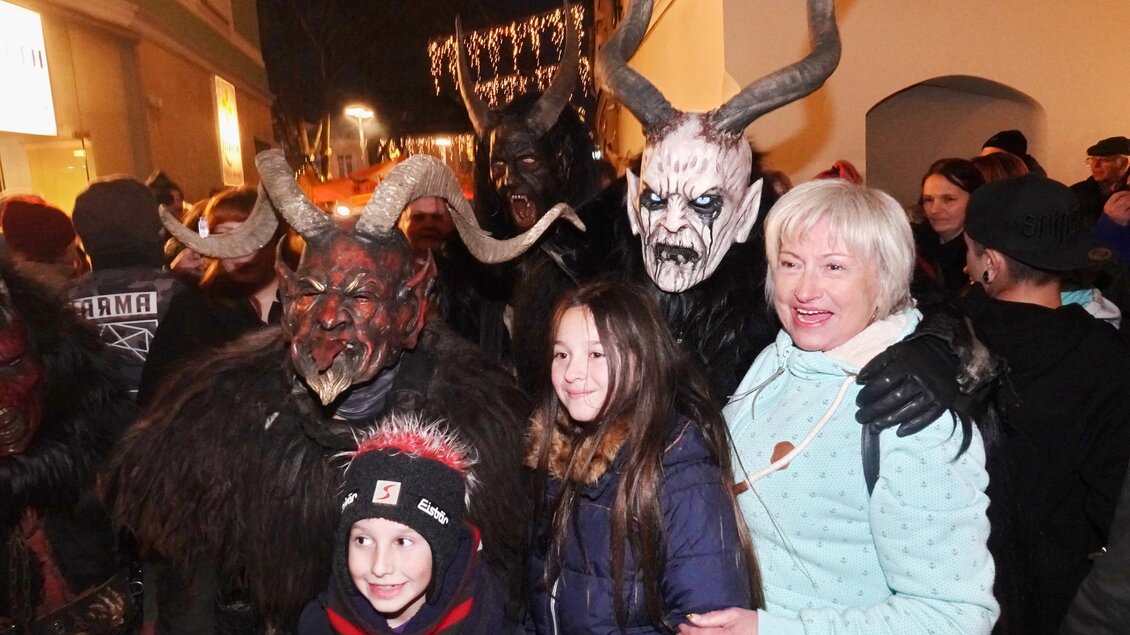Eine Gruppe von verkleideten Menschen mit Hörnern steht in einer Stadtstraße. Sie posieren lächelnd mit einer Familie, die ebenfalls im Bild ist.