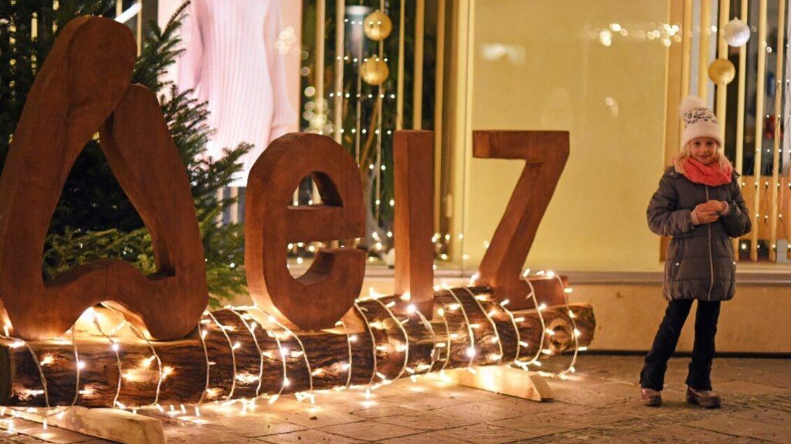 Ein kleines Mädchen steht neben einem großen, beleuchteten Holzschriftzug. Im Hintergrund sind dekorierte Weihnachtsbäume zu sehen. | © KK