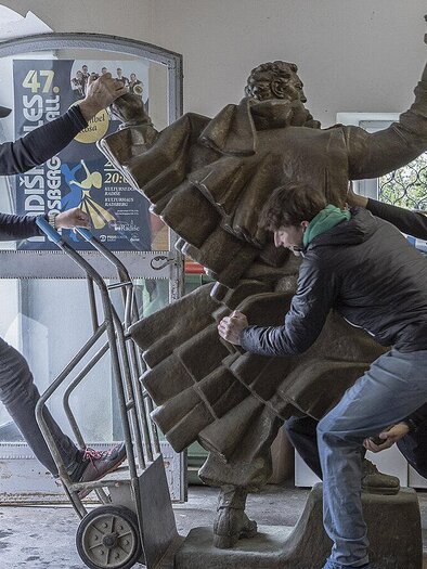 Drei Personen heben eine Statue und transportieren sie durch eine Tür. Die Umgebung sieht nach einem Lagerraum oder einer Werkstatt aus. | © Nicole Six und Paul Petritsch mit Jakob Holzer