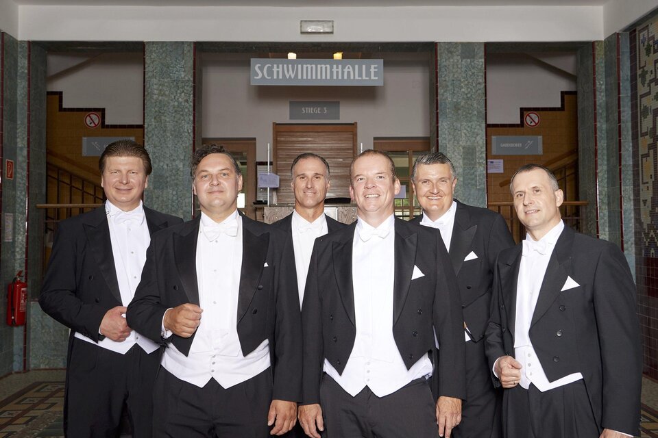 Sechs Männer in formeller Kleidung stehen vor einem Eingang zur Schwimmhalle. Sie lächeln in die Kamera und wirken freundlich. | © (c)KK