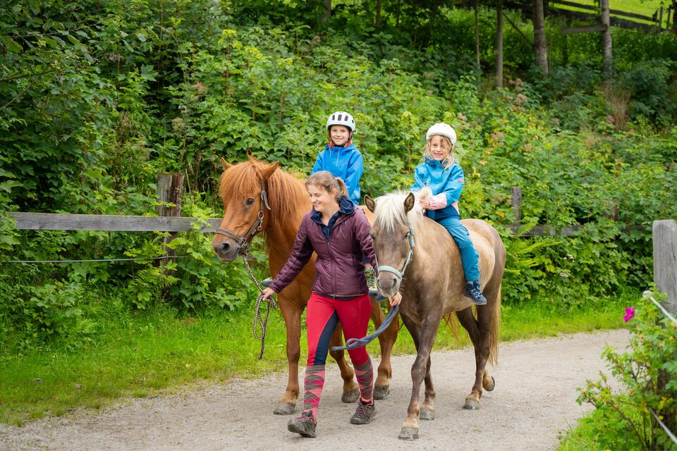 Eine Frau führt zwei Pferde auf einem Waldweg. Die Kinder auf den Pferden tragen Helme und lächeln. | © TVB/Laura Passenbrunner