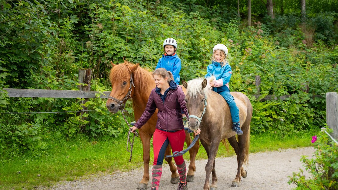 Eine Frau führt zwei Pferde auf einem Waldweg. Die Kinder auf den Pferden tragen Helme und lächeln. | © TVB/Laura Passenbrunner