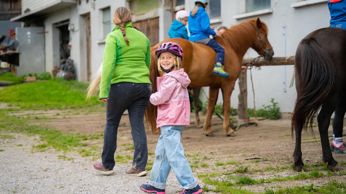 Ein Mädchen mit einem rosa Regenmantel und einem Helm lächelt, während sie auf einem Reiterhof läuft. Im Hintergrund sind andere Kinder auf Pferden zu sehen. | © TVB/Laura Passenbrunner