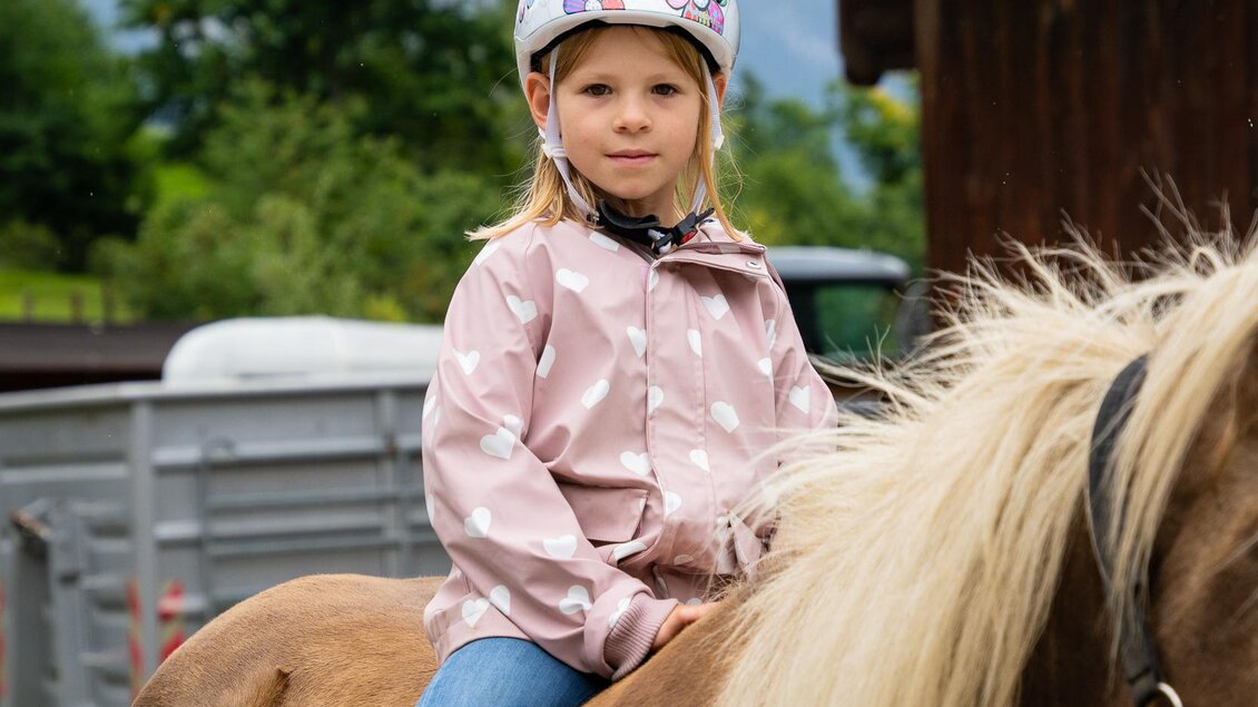 Ein Mädchen sitzt auf einem Pferd und trägt einen Helm. Im Hintergrund sind Bäume und eine Scheune zu sehen. | © TVB/Laura Passenbrunner
