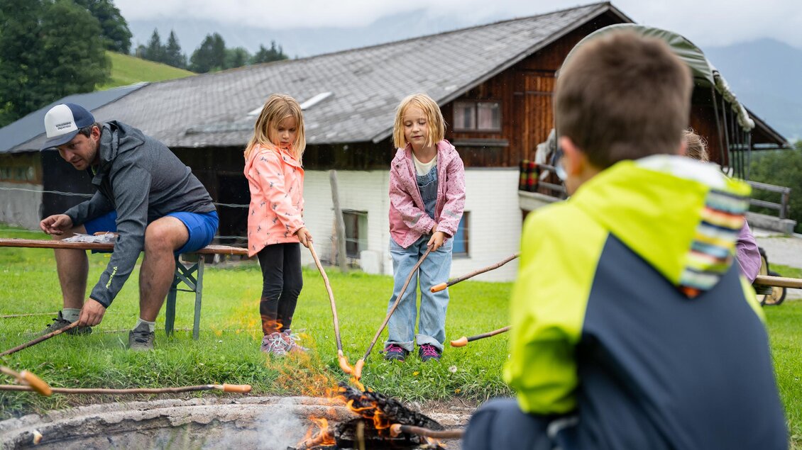 Ein paar Kinder grillen Würstchen über einem Lagerfeuer, während ein Erwachsener in der Nähe sitzt. Im Hintergrund ist ein Holzhaus und eine bewölkte Himmelslandschaft zu sehen. | © TVB/Laura Passenbrunner