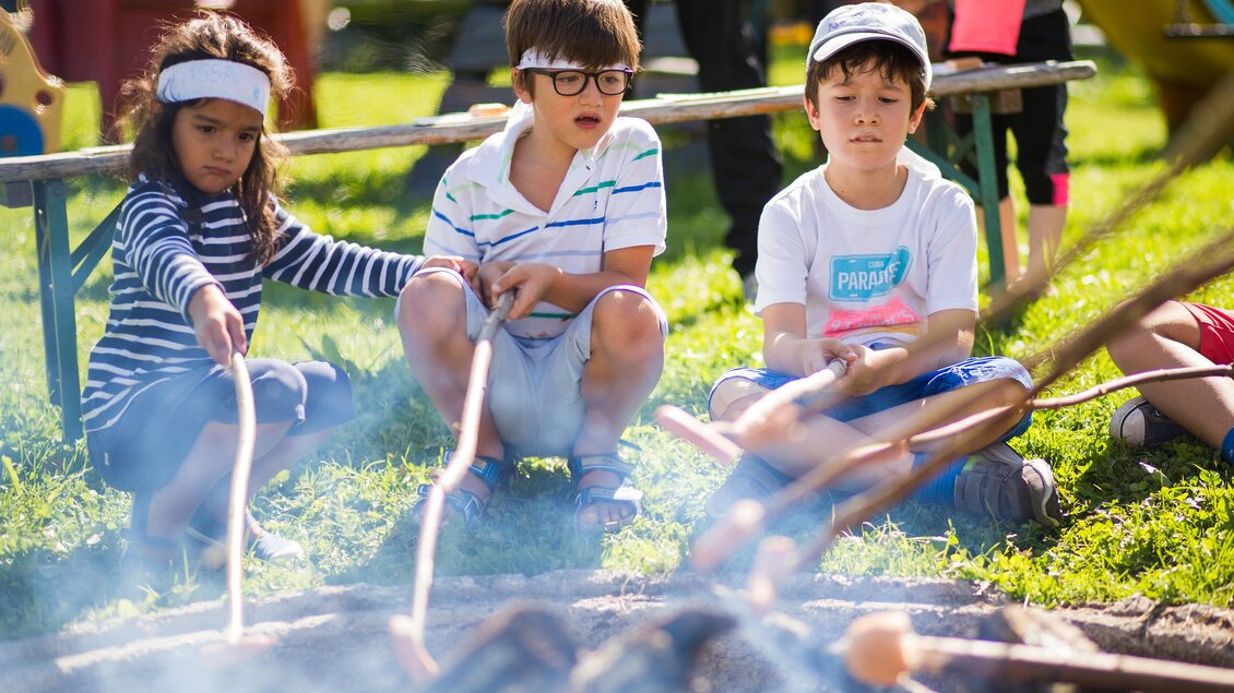Drei Kinder sitzen im Freien und grillen am Lagerfeuer. Sie halten Stöcke in der Hand und schauen auf das Feuer. | © Dominik Steiner