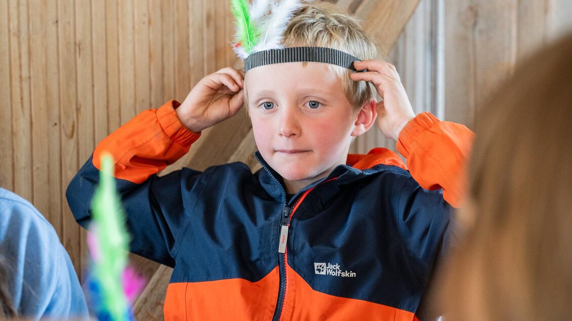 Ein Junge mit einem Stirnband und bunten Federn spielt in einem Raum. Er trägt eine orange und schwarze Jacke und schaut konzentriert. | © TVB/Laura Passenbrunner
