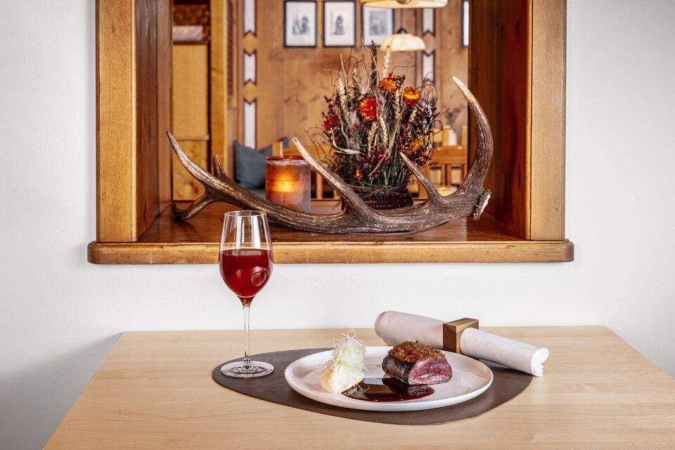 Ein elegantes Gericht auf einem Teller mit Fleisch, Beilage und einer süßen Creme. Im Hintergrund ein dekorativer Zierteil mit antler und ein Getränk in einem Weinglas. | © MONDI Hotels & Resorts