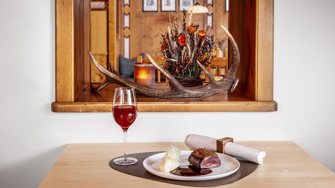 Ein elegantes Gericht auf einem Teller mit Fleisch, Beilage und einer süßen Creme. Im Hintergrund ein dekorativer Zierteil mit antler und ein Getränk in einem Weinglas. | © MONDI Hotels & Resorts