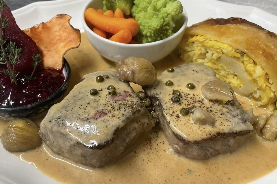 Ein köstliches Gericht mit zartem Fleisch in einer Sahnesauce, serviert mit buntem Gemüse und einer gefüllten Teigtasche. Auf dem Teller befinden sich außerdem eine Birne und eine kleine Schale mit Preiselbeermarmelade. | © Gasthof Steinmann