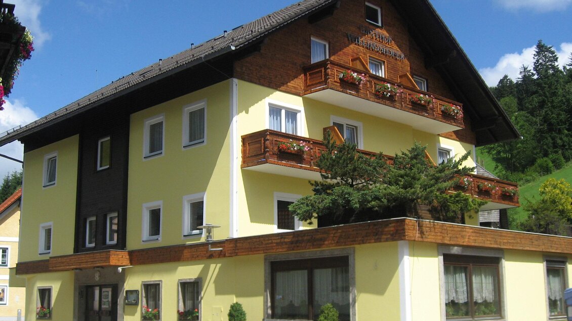 Ein schönes Gebäude mit gelber Fassade und Holzverkleidung. Es hat Balkone mit Pflanzen und liegt in einer grünen Umgebung. | © GH Willingshofer