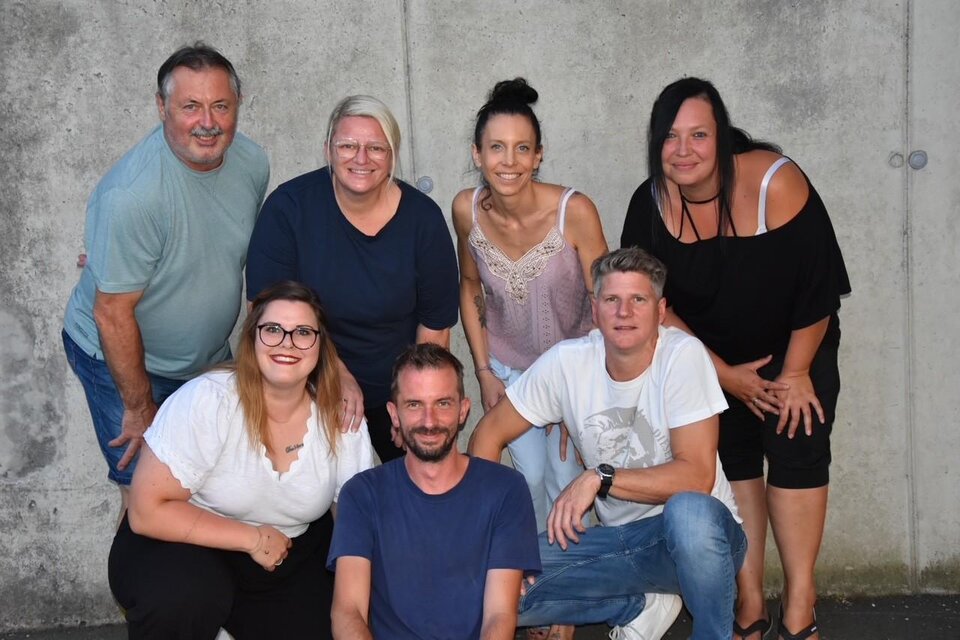 Eine Gruppe von sieben Personen posiert vor einer grauen Wand. Sie lächeln und scheinen Freude miteinander zu haben. | © Theatergruppe Wildrose