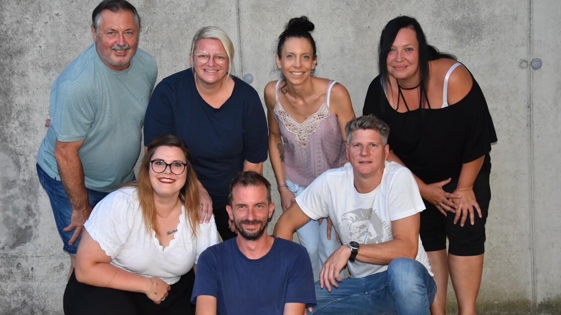 Eine Gruppe von sieben Personen posiert vor einer grauen Wand. Sie lächeln und scheinen Freude miteinander zu haben. | © Theatergruppe Wildrose