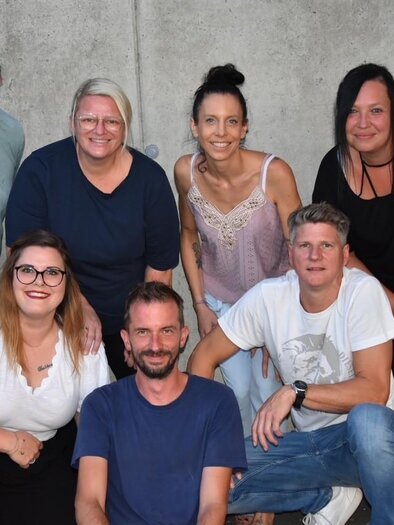 Eine Gruppe von sieben Personen posiert vor einer grauen Wand. Sie lächeln und scheinen Freude miteinander zu haben. | © Theatergruppe Wildrose