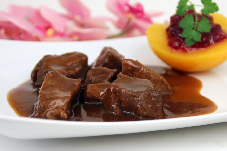 Ein Teller mit zartem Fleisch in einer schmackhaften Sauce, garniert mit frischen Kräutern. Neben dem Fleisch befindet sich ein Stück frischer Pfirsich mit einer Fruchtsauce. | © AdobeStock_38487448