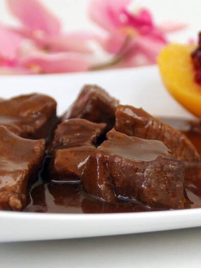 Ein Teller mit zartem Fleisch in einer schmackhaften Sauce, garniert mit frischen Kräutern. Neben dem Fleisch befindet sich ein Stück frischer Pfirsich mit einer Fruchtsauce. | © AdobeStock_38487448
