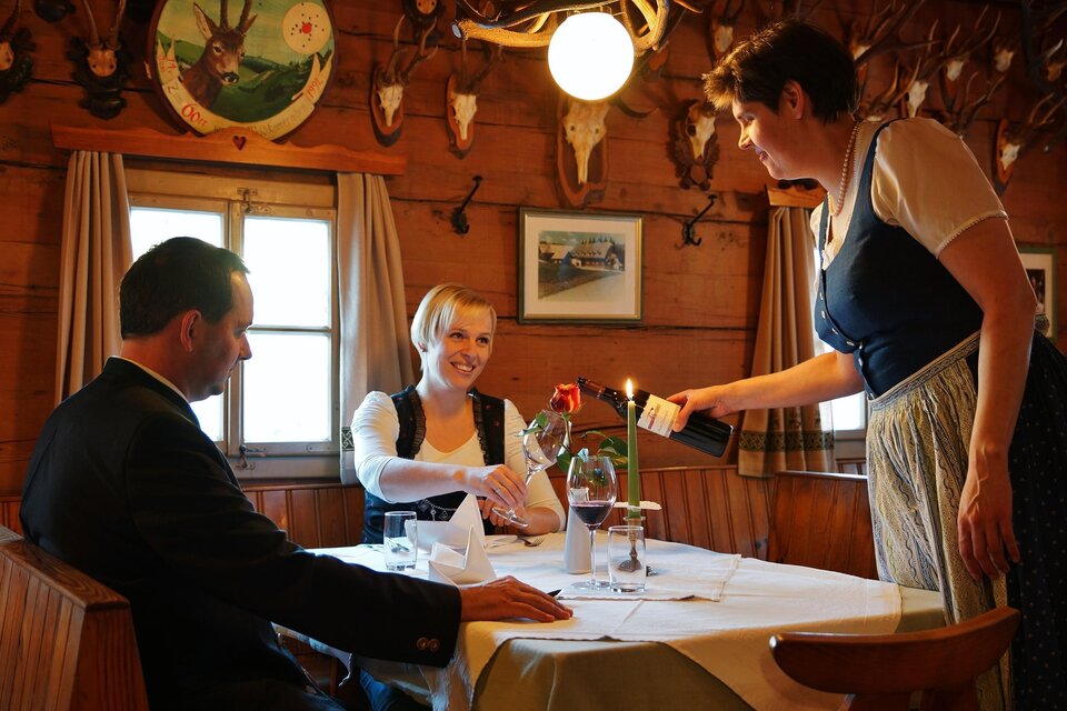 In einem gemütlichen Restaurant sitzt ein Paar am Tisch. Eine Kellnerin serviert eine Flasche Wein und eine Rose. | © Gasthof Unterberger