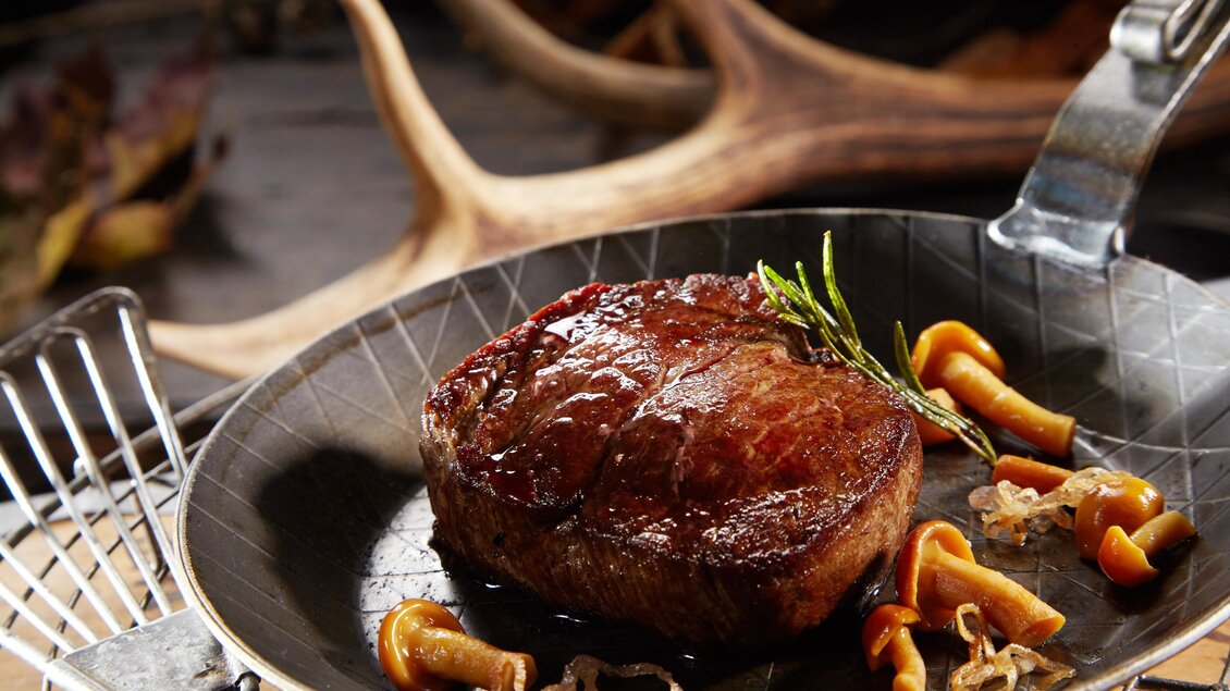 Ein saftiges Stück Fleisch auf einer Pfanne, garniert mit Kräutern und Pilzen. Im Hintergrund sind natürliche Elemente zu sehen, die eine rustikale Atmosphäre schaffen. | © AdobeStock