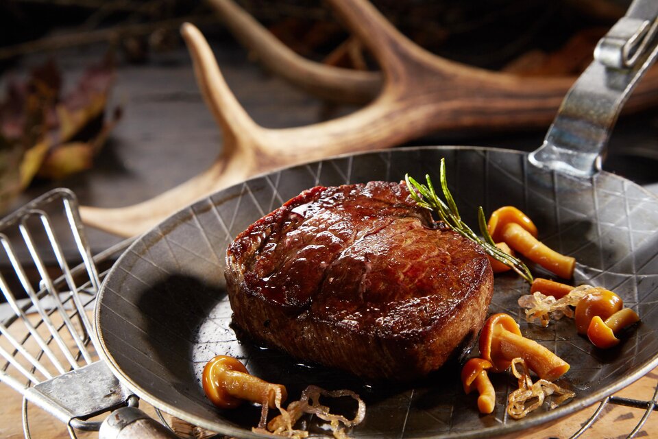 Ein saftiges Steak auf einer eleganten Pfanne, garniert mit frischen Kräutern und Pilzen. Im Hintergrund sind natürliche Elemente sichtbar, die eine rustikale Atmosphäre schaffen. | © AdobeStock