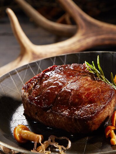 Egy szaftos steak egy elegáns serpenyőn, friss gyógynövényekkel és gombákkal díszítve. A háttérben természetes elemek láthatók, amelyek rusztikus légkört teremtenek. | © AdobeStock