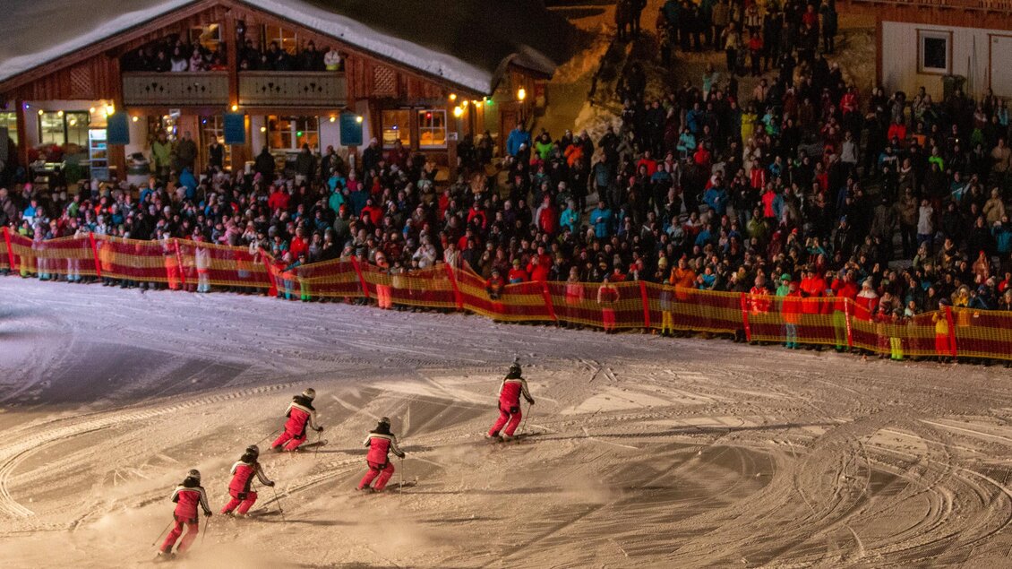 Eine lebhafte Wintersportveranstaltung mit Skifahrern in roten Anzügen, die auf der Piste fahren. Im Hintergrund ist eine große Menschenmenge und ein gemütliches Chalet zu sehen. | © Michael Simonlehner