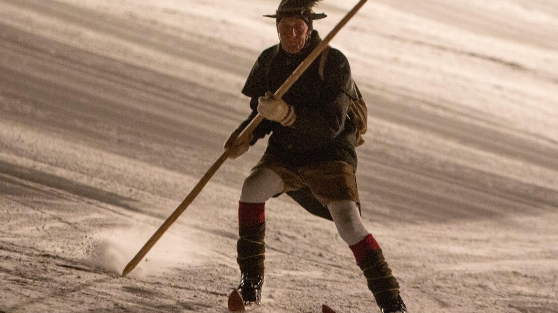 Ein Mann skiert in traditioneller Kleidung auf einer verschneiten Piste. Er hält einen Stock in der Hand und gleitet elegant über den Schnee. | © Michael Simonlehner