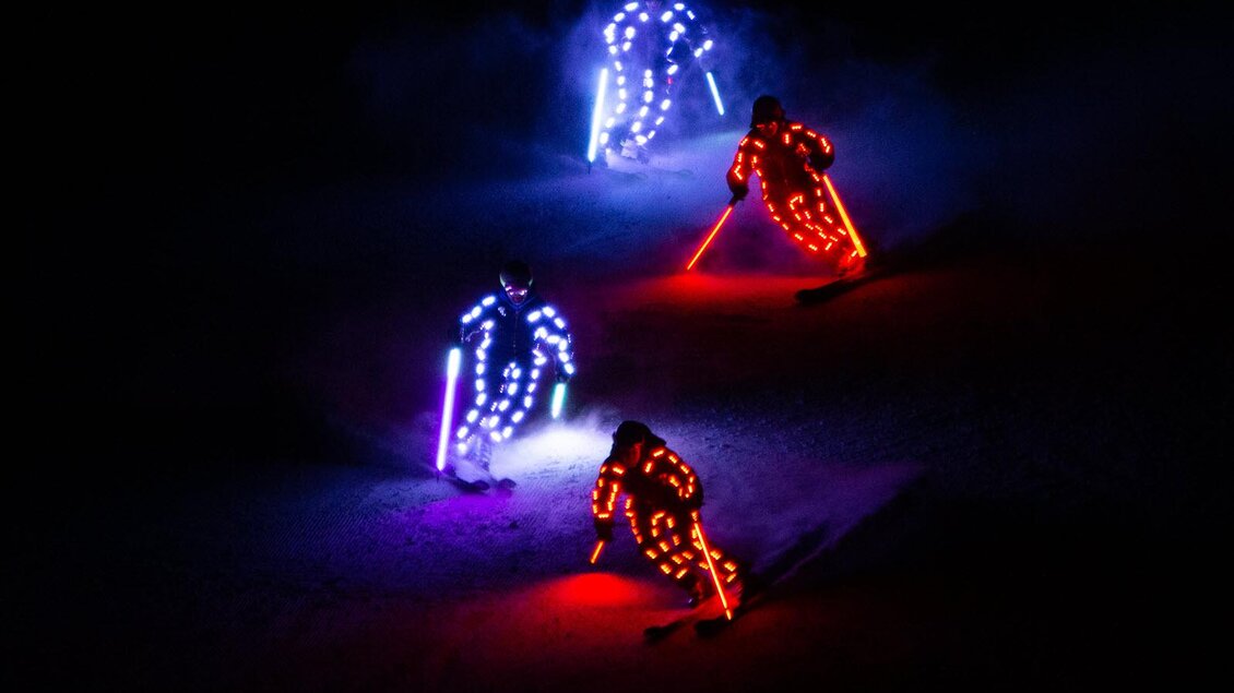 Drei Skifahrer mit leuchtenden Anzügen fahren durch den Schnee bei Nacht. Die bunten Lichter schaffen eine festliche Atmosphäre. | © Michael Simonlehner