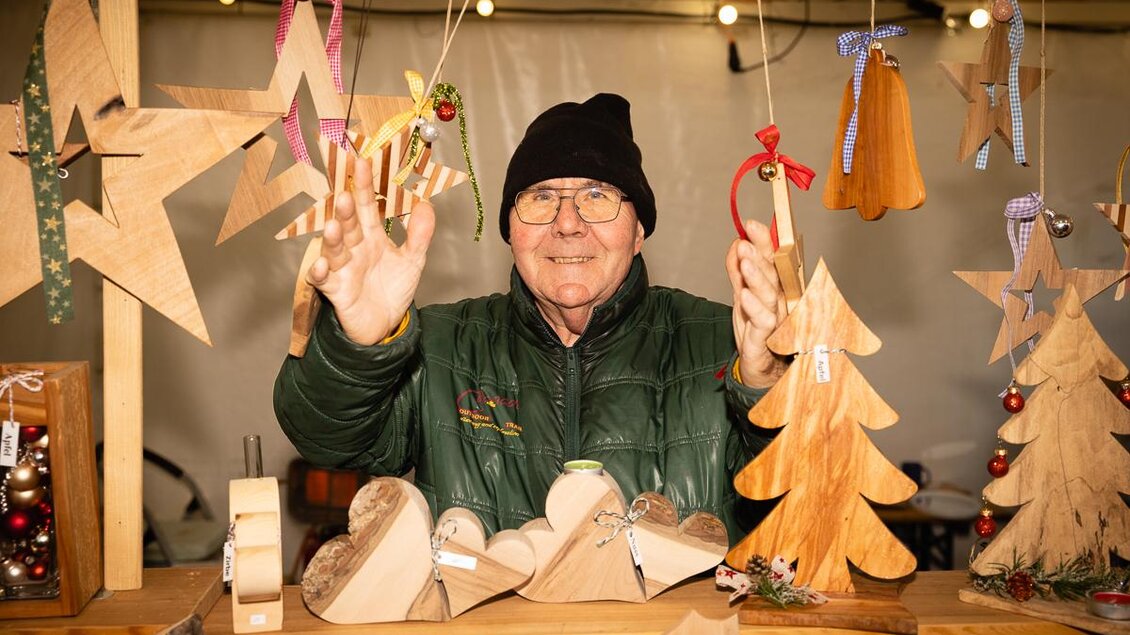 Ein älterer Mann steht an einem Marktstand mit handgefertigten Holzdekorationen. Um ihn herum hängen verschiedene Weihnachtsanhänger und Sterne. | ©  Roland Marx – copyright Grottenhof