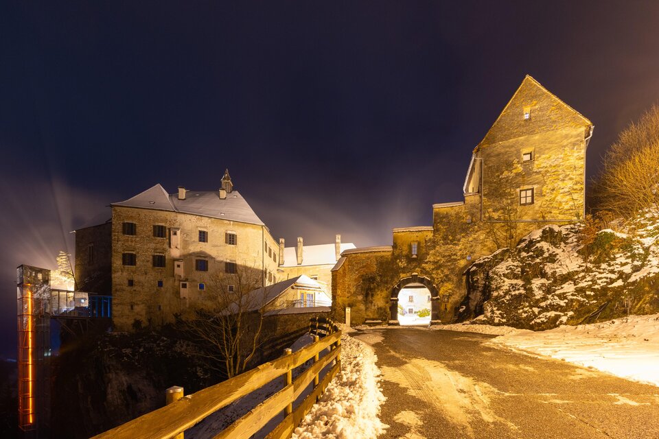 Eine alte Burg bei Nacht, umgeben von Schnee und sanften Lichtern. Die Straße führt zur Toröffnung der Burg und gibt einen faszinierenden Blick auf die Architektur. | © Region Graz-Harry Schiffer