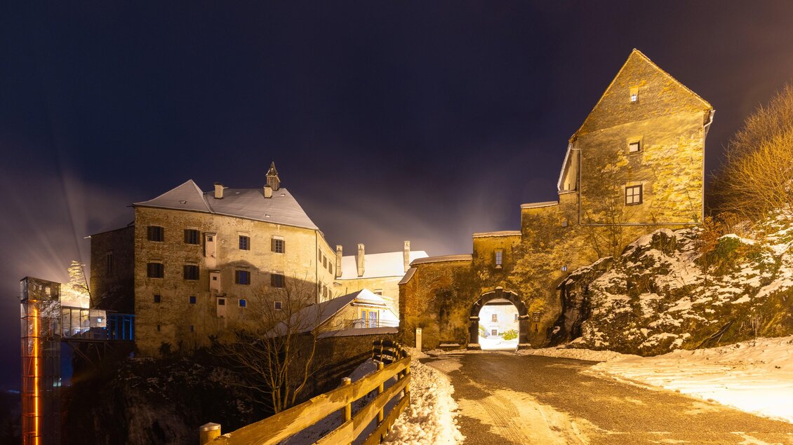 Eine alte Burg bei Nacht, umgeben von Schnee und sanften Lichtern. Die Straße führt zur Toröffnung der Burg und gibt einen faszinierenden Blick auf die Architektur. | © Region Graz-Harry Schiffer