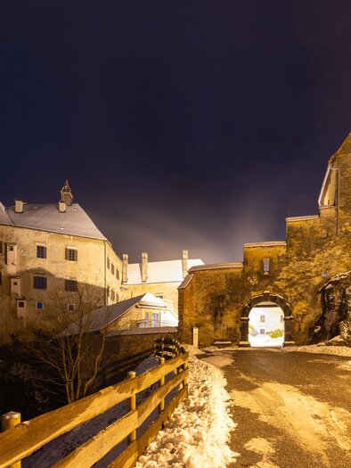 Eine alte Burg bei Nacht, umgeben von Schnee und sanften Lichtern. Die Straße führt zur Toröffnung der Burg und gibt einen faszinierenden Blick auf die Architektur. | © Region Graz-Harry Schiffer
