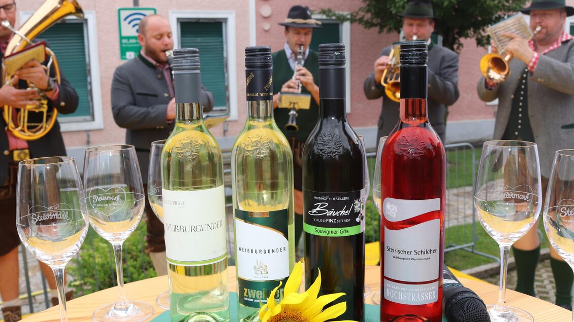 Eine Auswahl an Weinen steht auf einem Tisch, umgeben von Weingläsern und einer Sonnenblume. Im Hintergrund spielt eine Blaskapelle Musik. | © Gem. Mooskirchen-Robert Cescutti