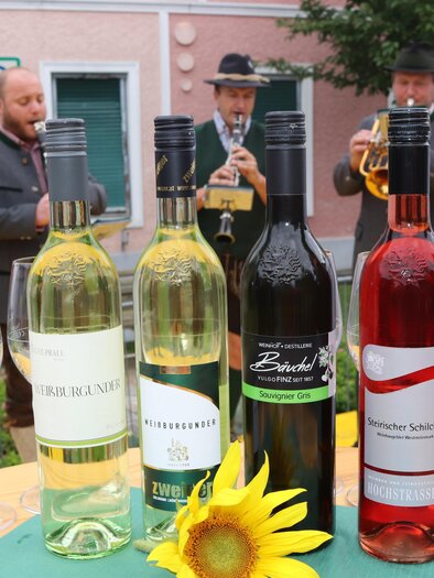 A selection of wines is on a table, surrounded by wine glasses and a sunflower. In the background, a brass band is playing music. | © Gem. Mooskirchen-Robert Cescutti
