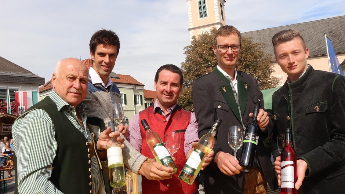 Eine Gruppe von fünf Männern in traditioneller Kleidung hält verschiedene Flaschen Wein und Gläser. Im Hintergrund sind Gebäude und Bäume zu sehen, was eine festliche Atmosphäre schafft. | © Gem. Mooskirchen-Robert Cescutti