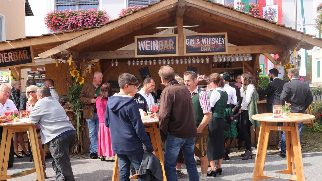 Ein gut besuchter Marktstand mit Gästen, die Getränke genießen. Einige Personen tragen traditionelle Trachten und die Umgebung ist festlich dekoriert. | © Gem. Mooskirchen-Robert Cescutti